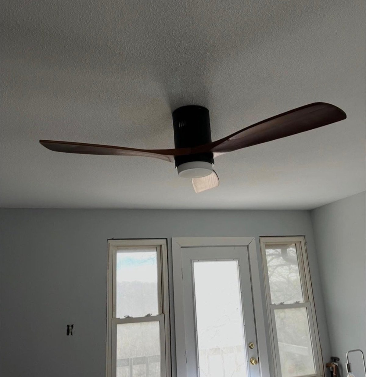 Modern black ceiling fan with three wooden blades installed on a white ceiling above glass doors