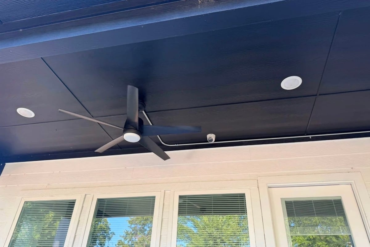 Modern black ceiling fan mounted on a dark blue covered porch ceiling above white-framed windows with green shutters