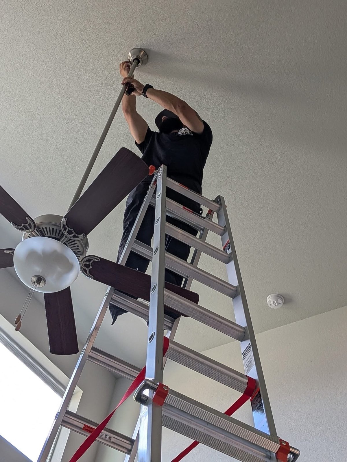 Person on an aluminum ladder reaching up to install a ceiling fan on a white ceiling