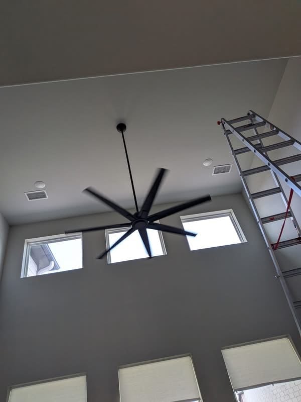 Large black ceiling fan mounted in a modern room with windows and a metal ladder visible on the right