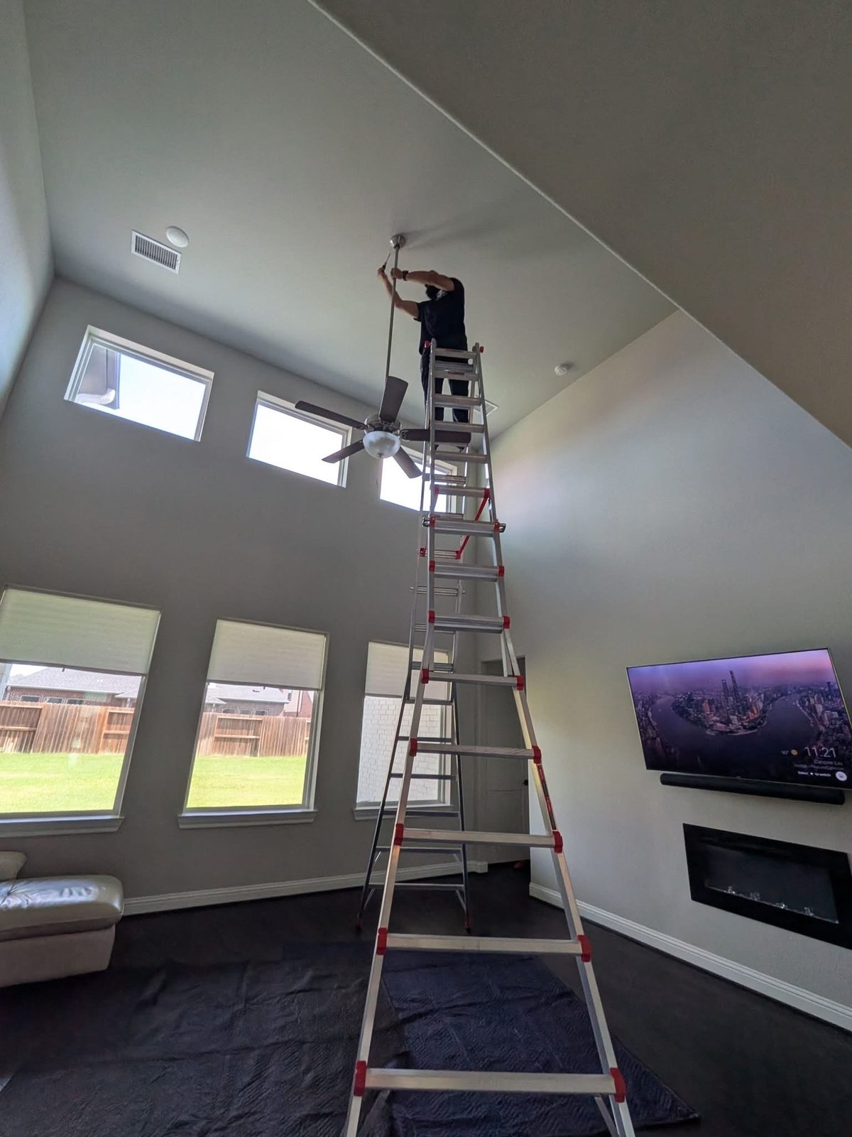 Worker on a tall ladder installing a ceiling fixture in a living room with tall windows and a TV in the background